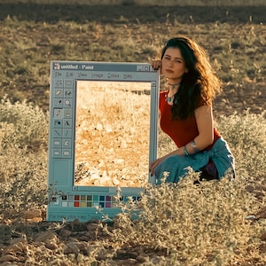 May include: A woman sits next to a large, rectangular frame resembling a computer window, labeled "untitled - Paint." The frame displays a sunlit field. The woman wears a red top and blue skirt, with jewelry. The scene is set outdoors.