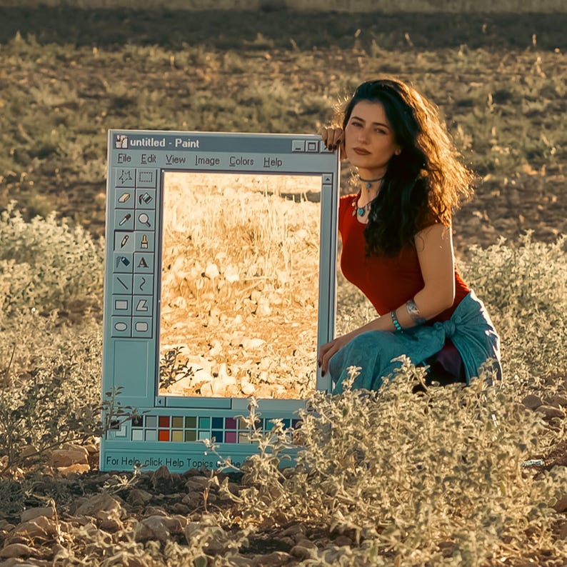 May include: A woman sits next to a large, rectangular frame resembling a computer window, labeled "untitled - Paint." The frame displays a sunlit field. The woman wears a red top and blue skirt, with jewelry. The scene is set outdoors.