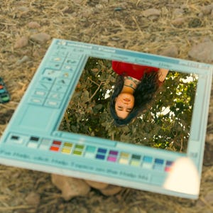 May include: A light blue, rectangular mirror designed to resemble a classic computer program interface. The mirror reflects a person wearing a red top and a turquoise necklace, set against a backdrop of green foliage. The mirror's frame features a toolbar and color palette.