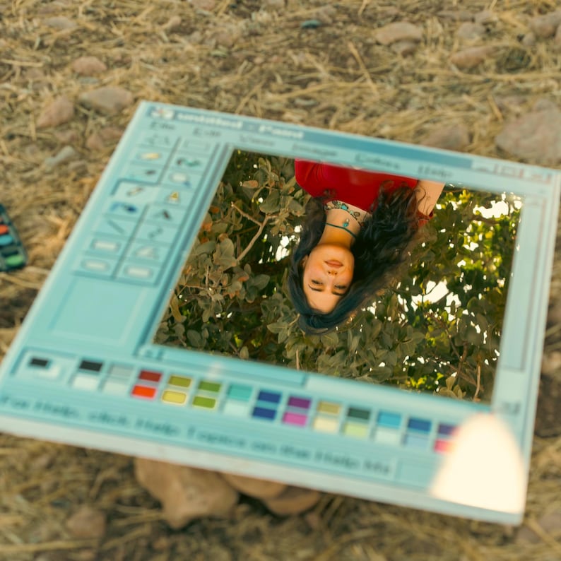May include: A light blue, rectangular mirror designed to resemble a classic computer program interface. The mirror reflects a person wearing a red top and a turquoise necklace, set against a backdrop of green foliage. The mirror's frame features a toolbar and color palette.