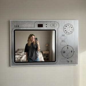 Puede incluir: Un espejo de pared rectangular diseñado para parecerse a la parte posterior de una cámara digital. El marco presenta un acabado gris metálico con controles de cámara impresos, incluyendo un dial de modo, botones de menú e iconos de navegación.