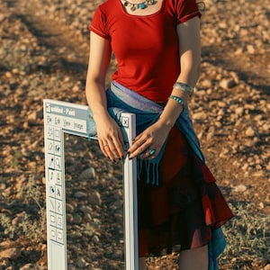 May include: A white framed prop with the text "untitled - Paint" and a toolbar. The person is wearing a red top, a patterned skirt, and a blue scarf. The background is a dry, rocky landscape.