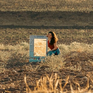 May include: A woman kneels beside a vintage computer monitor displaying a landscape. The monitor, with the Paint program interface visible, reflects the golden-hued field. The woman wears a red top and blue jeans, blending with the natural setting.