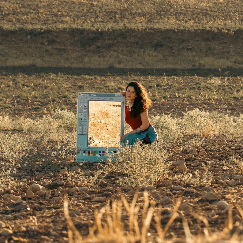 May include: A woman kneels beside a vintage computer monitor displaying a landscape. The monitor, with the Paint program interface visible, reflects the golden-hued field. The woman wears a red top and blue jeans, blending with the natural setting.