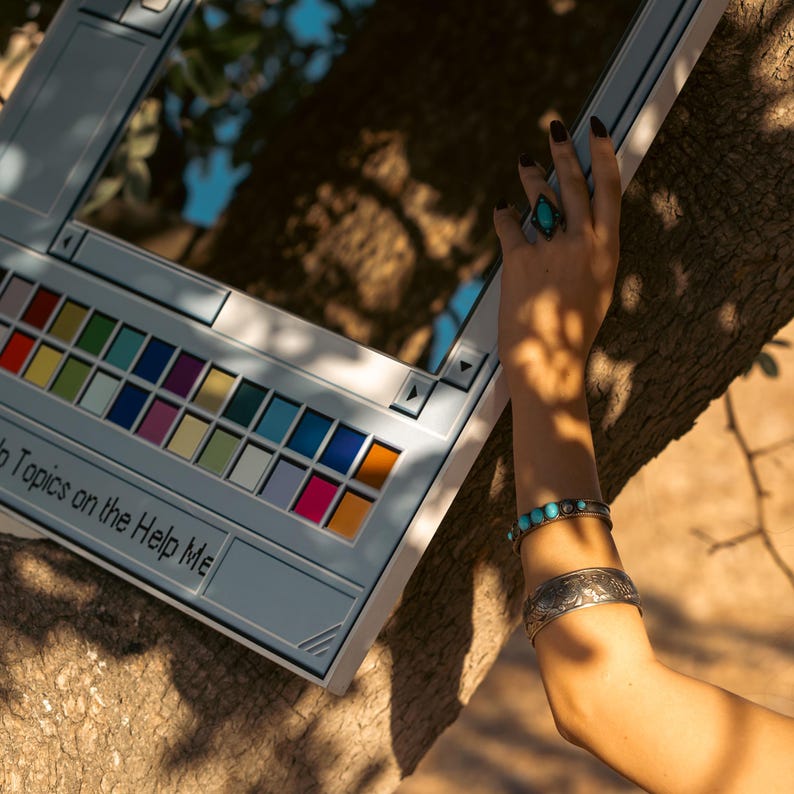 May include: A light blue mirror in the shape of a computer window, with a colorful palette of paint colors. The text "Help Me" is visible. The mirror is resting on a tree branch.