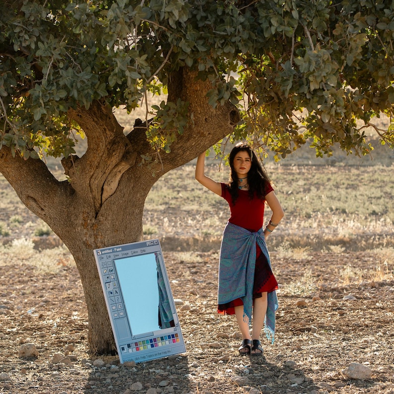 May include: A young person stands near a large tree, wearing a red top and a blue wrap skirt. A large, rectangular mirror with a Windows Paint interface design leans against the tree trunk. The mirror reflects the sky.