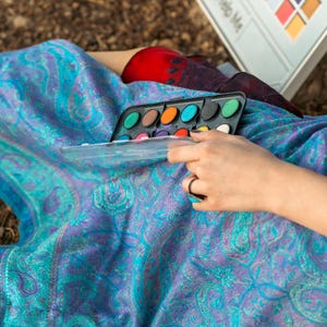 May include: A watercolor paint set with a clear lid, featuring various colors, held by a person. The person is wearing a ring with a turquoise stone. The background includes a patterned blue and purple fabric.
