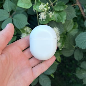 Puede incluir: Un pequeño recipiente blanco de forma ovalada y superficie lisa. El recipiente se sostiene en una mano sobre un fondo de hojas verdes y flores blancas. El recipiente parece ser un producto cosmético o de cuidado personal.