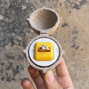 May include: A gold ring with a large, clear gemstone is displayed in an open, yellow ring box. The ring box is inside a round, beige, egg-shaped container. The ring is the focal point, showcasing its brilliance.