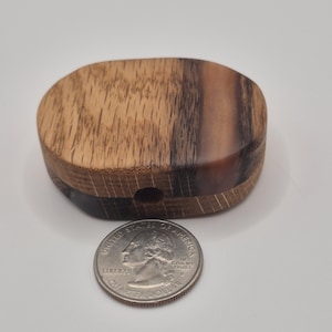 May include: A small, wooden box with a rounded shape, featuring a mix of light and dark wood grains. A U.S. quarter coin is placed next to the box for size comparison. The box has a small hole on one side.