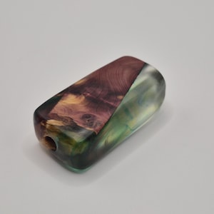 May include: A handcrafted, rectangular wooden bead with a hole. The bead features a combination of rich, natural wood tones and translucent green and blue resin. The wood grain is visible, adding to the unique, artistic appeal of the bead.