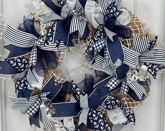 May include: A nautical-themed wreath with a burlap base, adorned with navy blue and white ribbons. The ribbons feature anchor, striped, and gingham patterns. White mesh and curly accents add texture to the circular design.