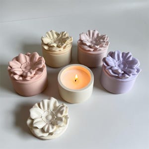 Peut inclure: Plusieurs bougeoirs en forme de fleurs aux couleurs pastel, dont rose, lavande et crème. Une bougie est allumée, entourée de contenants décoratifs. Les couvercles sont en forme de fleurs détaillées.