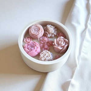 Peut inclure: Un bougeoir rond blanc rempli de bougies en forme de fleurs roses et mauves. Les formes de bougies comprennent des roses et d'autres motifs floraux détaillés. L'arrangement est posé sur une surface blanche avec un tissu blanc en arrière-plan.