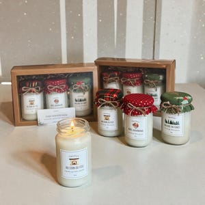 May include: A lit candle in a glass jar with a label that reads "Au Coin Du Feu". Several other glass jars with decorative fabric lids are in the background. Some jars are in a wooden box. The jars are filled with white wax.