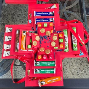 May include: A red explosion box with various treats and gifts, including chocolate bars, candies, and gold-colored coins. The box is decorated with the number 70 and the British Airways logo, suggesting a birthday theme. Red ribbon adds a decorative touch.