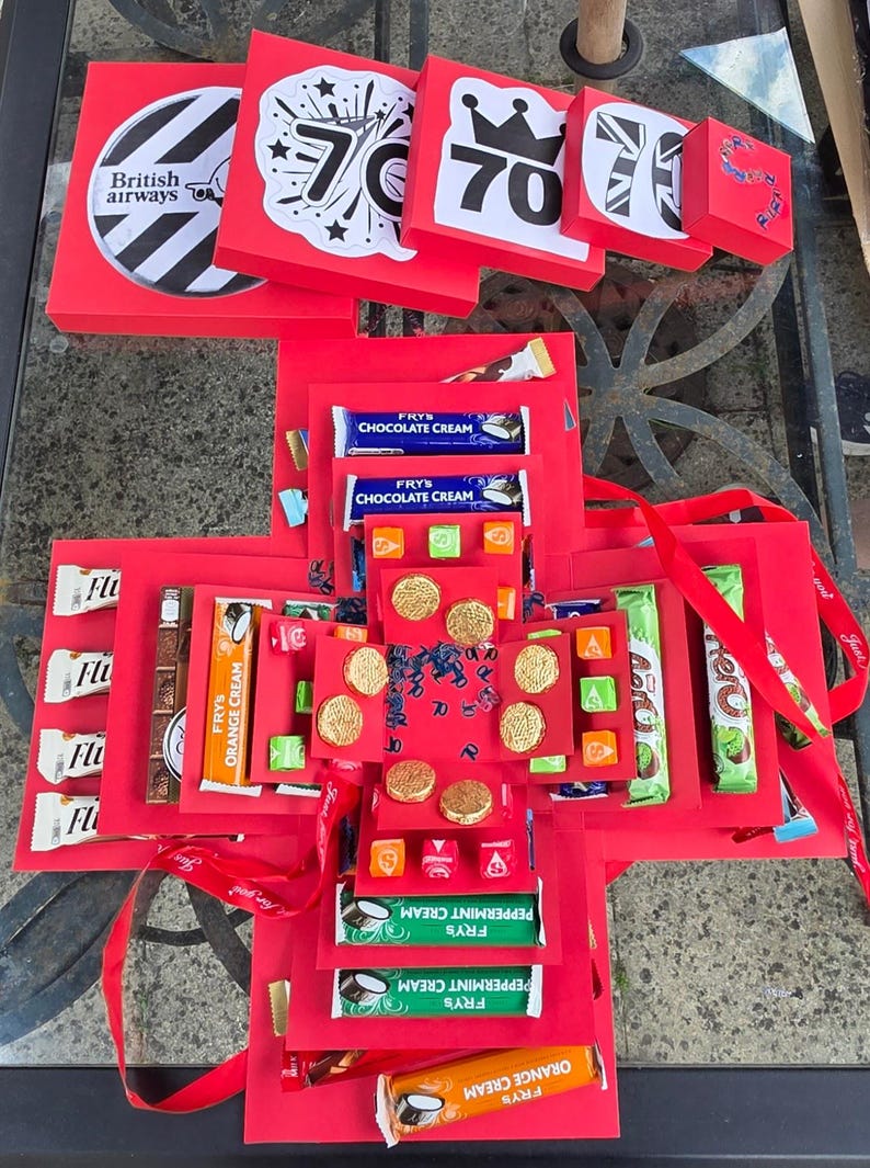 May include: A red explosion box with various treats and gifts, including chocolate bars, candies, and gold-colored coins. The box is decorated with the number 70 and the British Airways logo, suggesting a birthday theme. Red ribbon adds a decorative touch.