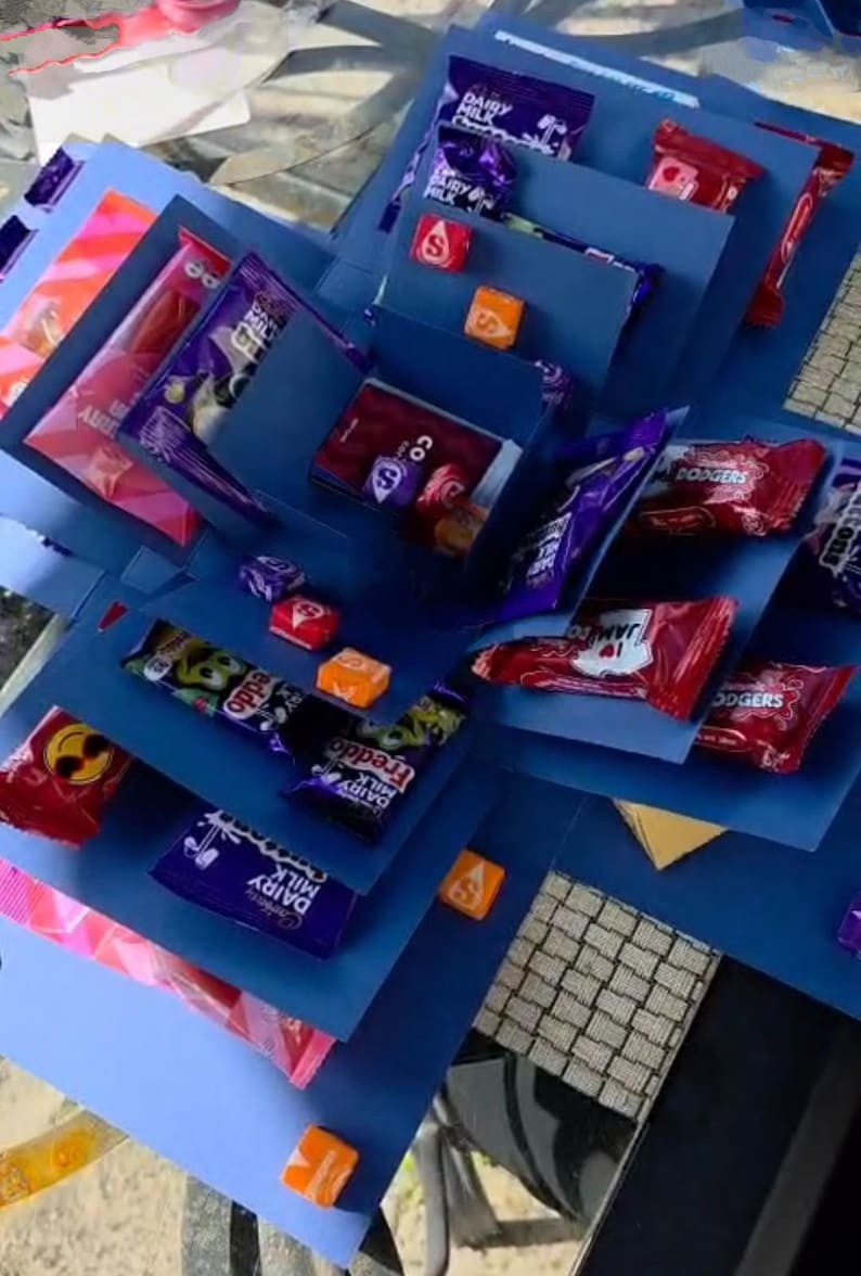 May include: A blue explosion box filled with various wrapped candies and chocolates. The box is open, revealing layers of treats, including Dairy Milk, Freddo, and other brands. Orange and red candies are also visible.