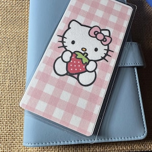 Puede incluir: Una agenda azul claro con una cubierta de plástico transparente. La cubierta tiene un patrón de cuadros rosas y un gráfico de Hello Kitty sosteniendo una fresa. La agenda tiene un cierre a presión.