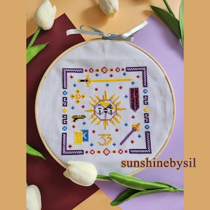 Puede incluir: Bordado de punto de cruz en un aro de madera con un diseño de sol con una cara, espada, escudo y otros símbolos. El diseño está enmarcado en un borde púrpura con detalles en rojo, azul y amarillo. El texto "sunshinebysil" está en la parte inferior.