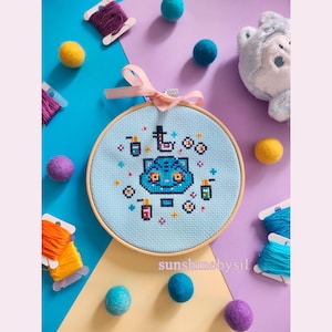 May include: A cross-stitch embroidery hoop featuring a blue cat design with a top hat and various colorful elements. The text "sunshinebysil" is visible. The hoop is surrounded by colorful yarn, felt balls, and a plush toy.