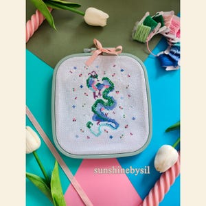 Puede incluir: Un bordado de punto de cruz con un diseño de dragón verde y azul estilizado sobre tela blanca, en un aro cuadrado verde claro. El diseño incluye pequeñas puntadas de cruz azules y rosas. Una cinta rosa está atada en la parte superior. El texto "sunshinebysil" está en la parte inferior.