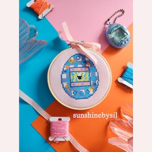 May include: A cross-stitch embroidery hoop featuring a pixelated design in blue, yellow, and pink, resembling a retro digital pet. The hoop is tied with a pink ribbon. The background includes spools of thread and a vintage digital pet.