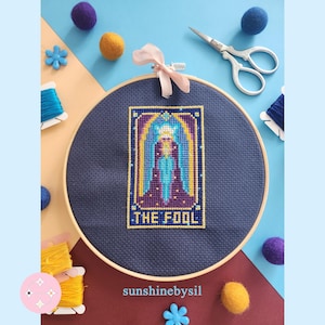 Puede incluir: Un bordado de punto de cruz de la carta del tarot "El Loco", en un aro de madera. El diseño presenta una figura en morado y azul, con detalles dorados, sobre un fondo azul marino. El texto "THE FOOL" está en la parte inferior.
