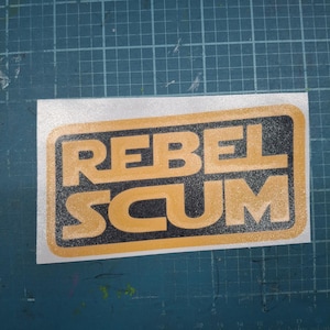 May include: A rectangular sticker with the words "REBEL SCUM" in gold block letters. The text is set against a black background within a gold rectangular border. The sticker is on a green cutting mat.