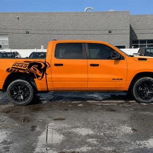 Puede incluir: Una camioneta pickup de color naranja brillante con cabina doble y una calcomanía gráfica negra en el panel lateral trasero. El diseño muestra un bloque de motor estilizado con un emblema de cabeza de carnero. Tiene ruedas, manijas y molduras negras.