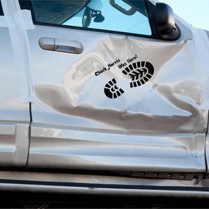 May include: A silver vehicle with a heavily dented door, taped with clear plastic. Black text and a footprint graphic reading "Chuck Norris Was Here!" are visible on the door.