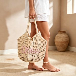 May include: A cream-coloured crochet tote bag featuring an open-knit mesh design and two shoulder straps. The front of the bag displays the name Chloe in raised, light pink cursive lettering. The bag is constructed from a textured yarn material.