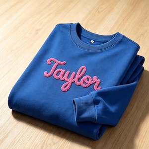 May include: A royal blue crewneck sweatshirt featuring the name Taylor written in pink, textured, fuzzy chenille-style script lettering across the chest. The garment has long sleeves with ribbed cuffs and a ribbed neckline, showcasing a soft fabric.