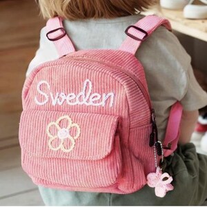 Può includere: Uno zaino in velluto a coste rosa con la parola Sweden ricamata in corsivo bianco sulla parte superiore. La tasca frontale presenta un disegno a fiore giallo cucito e un piccolo ciondolo a farfalla all'uncinetto rosa pende dalla zip.