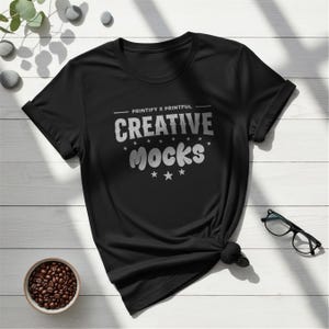 以下が含まれることがあります： 「CREATIVE MOCKS」の文字が銀色の少し擦れたフォントでプリントされた黒いTシャツ。Tシャツはクルーネックで半袖です。「PRINTIFY X PRINTFUL」の文字がメインテキストの上にあります。コーヒー豆の小さなボウルとメガネも表示されています。