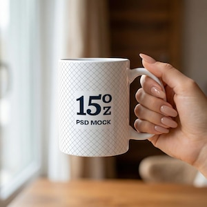 May include: White ceramic mug with a grid pattern and the text "15 oz PSD MOCK" in black. The mug is held by a hand with manicured nails. The background is blurred, showing a window and wooden table.