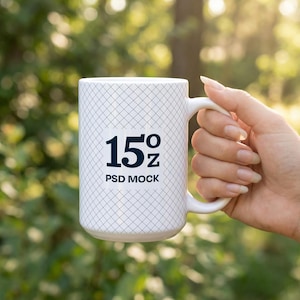 May include: White ceramic mug with a grid pattern, held by a hand. The mug features the text "15 oz PSD MOCK" in black. The background is a blurred view of green foliage and sunlight.