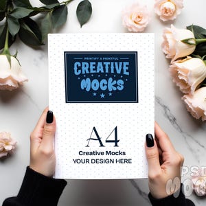 Puede incluir: Cuaderno tamaño A4 con una cubierta blanca punteada, sostenido por manos con esmalte de uñas negro. La portada presenta el texto "PRINTIFY X PRINTFUL" y "CREATIVE MOCKS" en azul, con estrellas. La parte inferior dice "YOUR DESIGN HERE".