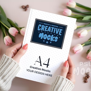 May include: A white A4 notebook with a blue and white design that reads "CREATIVE MOCKS". The notebook is held in front of a soft pink background with pink tulips and small brown decorative elements.