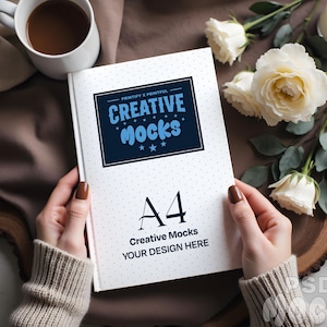 Peut inclure: Un cahier A4 avec le texte "Creative Mocks" dans un encadré bleu marine, tenu par une personne. Le cahier a une couverture blanche avec de petits pois gris. Une tasse de café et des roses blanches sont en arrière-plan.