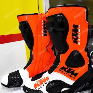 Puede incluir: Un par de botas de carreras para motocicleta de caña alta en naranja brillante, blanco y negro. Las botas presentan el logotipo de KTM Racing en texto negro y naranja, espinilleras negras, punteras reforzadas y paneles flexibles en el tobillo.