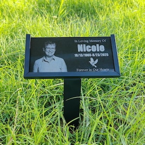 May include: A black memorial plaque with the name "Nicole" and dates 10/13/1965-8/23/2025, featuring a photo and the text "In Loving Memory Of" and "Forever in Our Hearts." The plaque is set in green grass.
