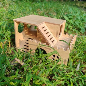 May include: A wooden hamster house with multiple levels, including a rooftop, a ramp, and a lower level with a decorative arched window. The house is made of light-colored wood and is surrounded by green grass.