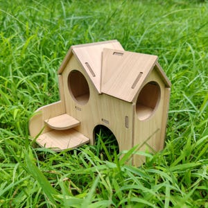 May include: A wooden pet house with a house-like design, featuring a sloped roof, round entry holes, and a small set of steps. The structure is made of light-colored wood and sits in a bed of green grass.