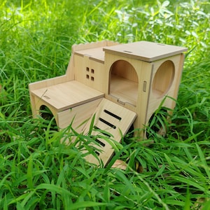 May include: A wooden hamster house with multiple levels, including a ramp, windows, and a small balcony. The house is made of light-colored wood and sits in a bed of green grass.
