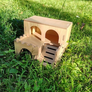 May include: A light-colored wooden hamster house with a castle-like design. The structure features arched doorways, a small staircase, and a crenelated roofline. The house is set in a bed of green grass.
