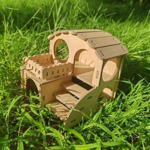 Könnte beinhalten: Ein mehrstöckiges Hamsterhaus aus Holz mit Balkon, Treppe und kreisförmigen Öffnungen. Es besteht aus hellem Holz mit dunkleren Holzakzenten auf dem Dach. Das Haus steht in grünem Gras.