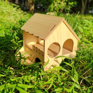 Puede incluir: Una pequeña casa de madera para mascotas, con un acabado marrón claro. La casa presenta un borde de techo festoneado, aberturas arqueadas y un pequeño balcón. Diseñada para mascotas pequeñas, está situada en hierba verde.