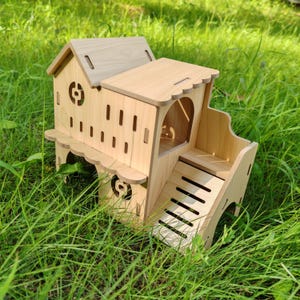 May include: A light-colored wooden hamster house with a multi-level design. The house features a small staircase, decorative cutouts, and a sloped roof. The structure is set against a backdrop of green grass.