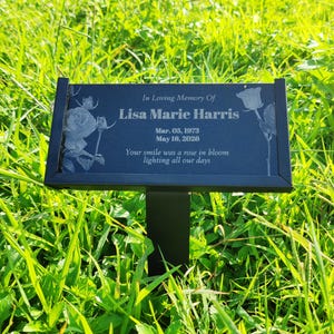 Peut inclure: Plaque commémorative noire avec l'inscription "In Loving Memory Of Lisa Marie Harris" et des dates. La plaque présente des illustrations de roses gravées et est montée sur un piquet en métal noir, dans une zone herbeuse.